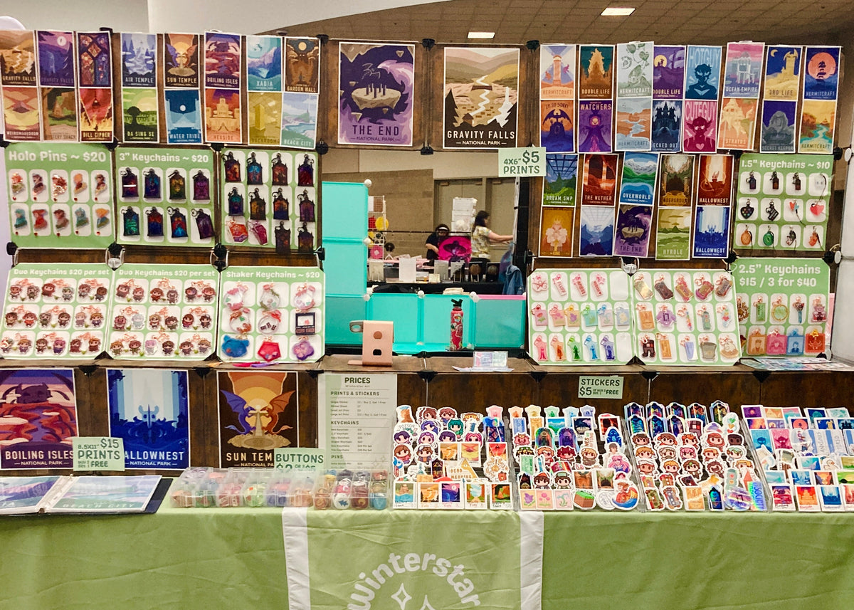 Art display with various artworks on a table and wall, featuring a green tablecloth and 'Winterstar Art' banner.